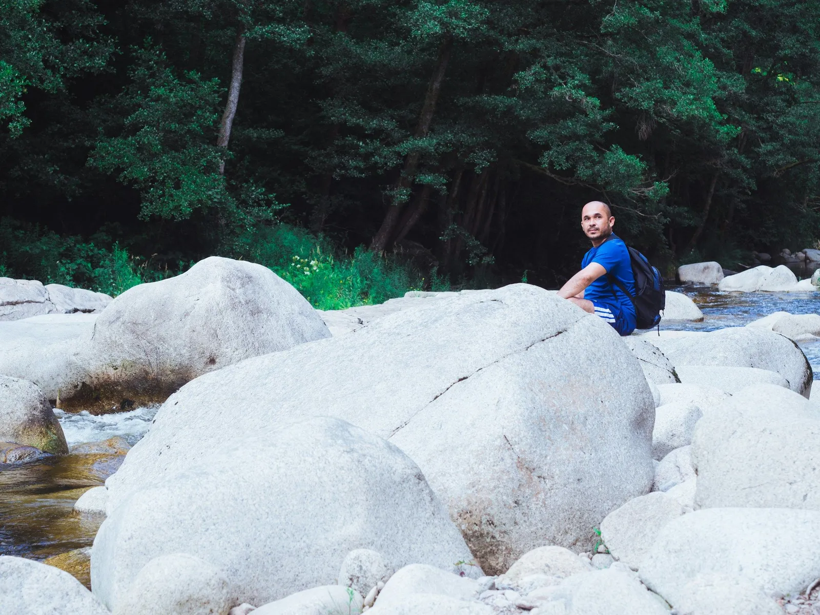Man in blue shirt with backpack sitting on large white boulders beside a forest stream, looking back over his shoulder.