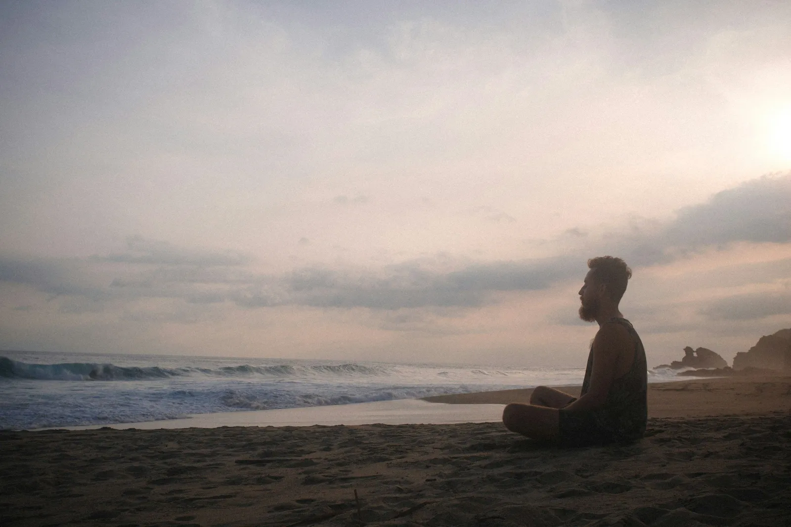 A bearded man sits cross-legged in meditation on a sandy beach at dusk, facing breaking waves under a pastel cloudy sky.