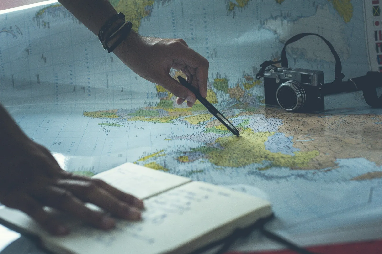 Top-down shot of hands planning a trip with a paper world map, an open notebook with pen, and a vintage camera on a wooden surface.