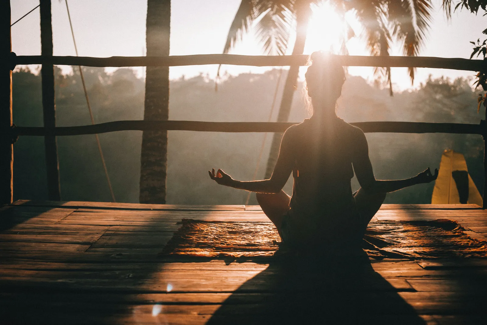 Lotus pose silhouette on a wooden deck at sunrise with palm trees and jungle. The classic budget-retreat moment: an outdoor practice space that costs nothing extra.