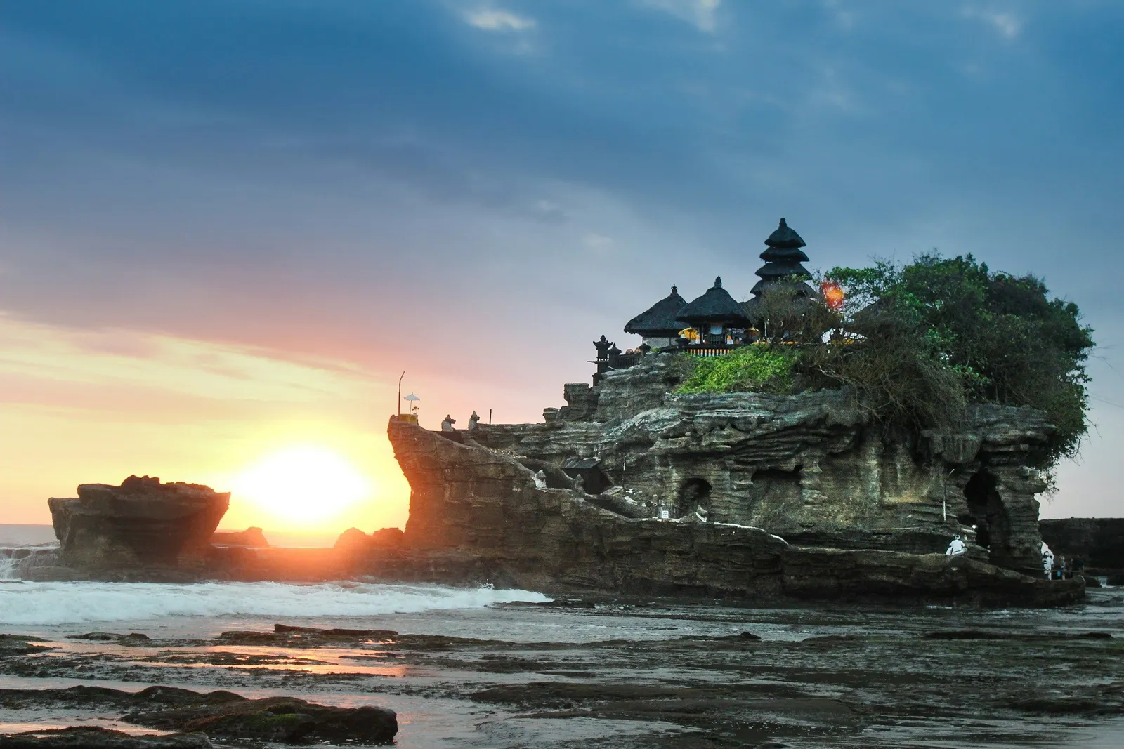 Tanah Lot temple perched on a rocky outcrop in Bali at sunset, the sun low on the horizon and waves washing the rocks below. Warm orange and blue light.
