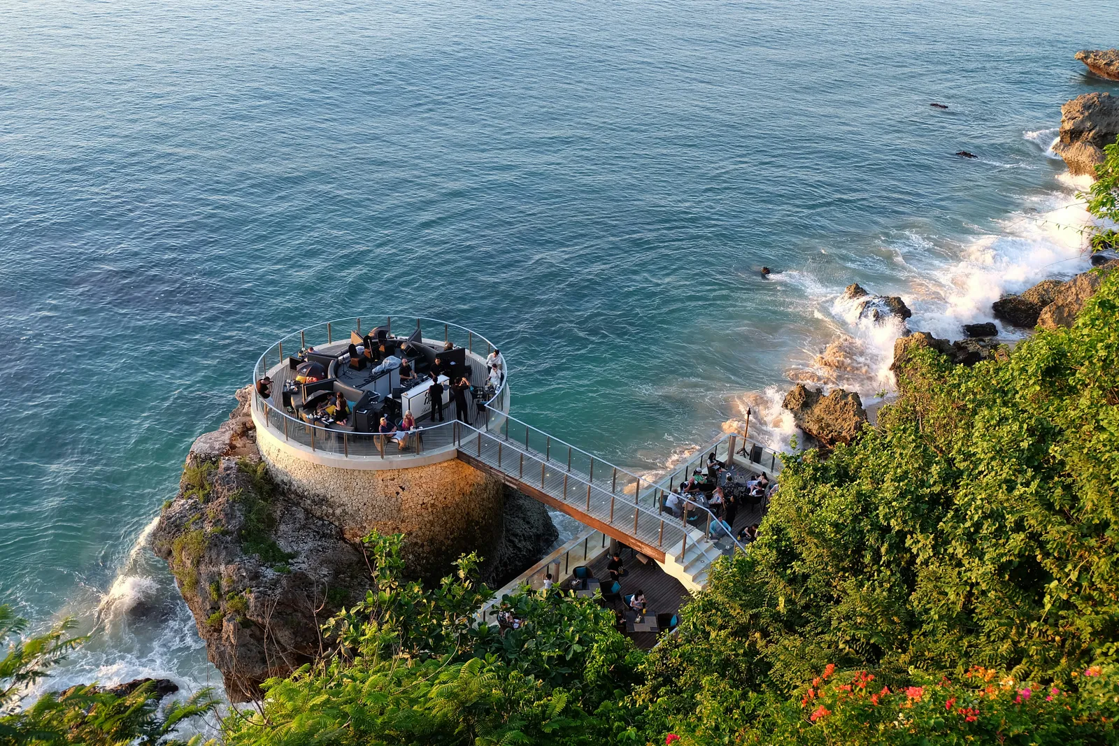 Aerial view of a clifftop circular dining platform overlooking turquoise sea with waves crashing onto rocks below, Bali.
