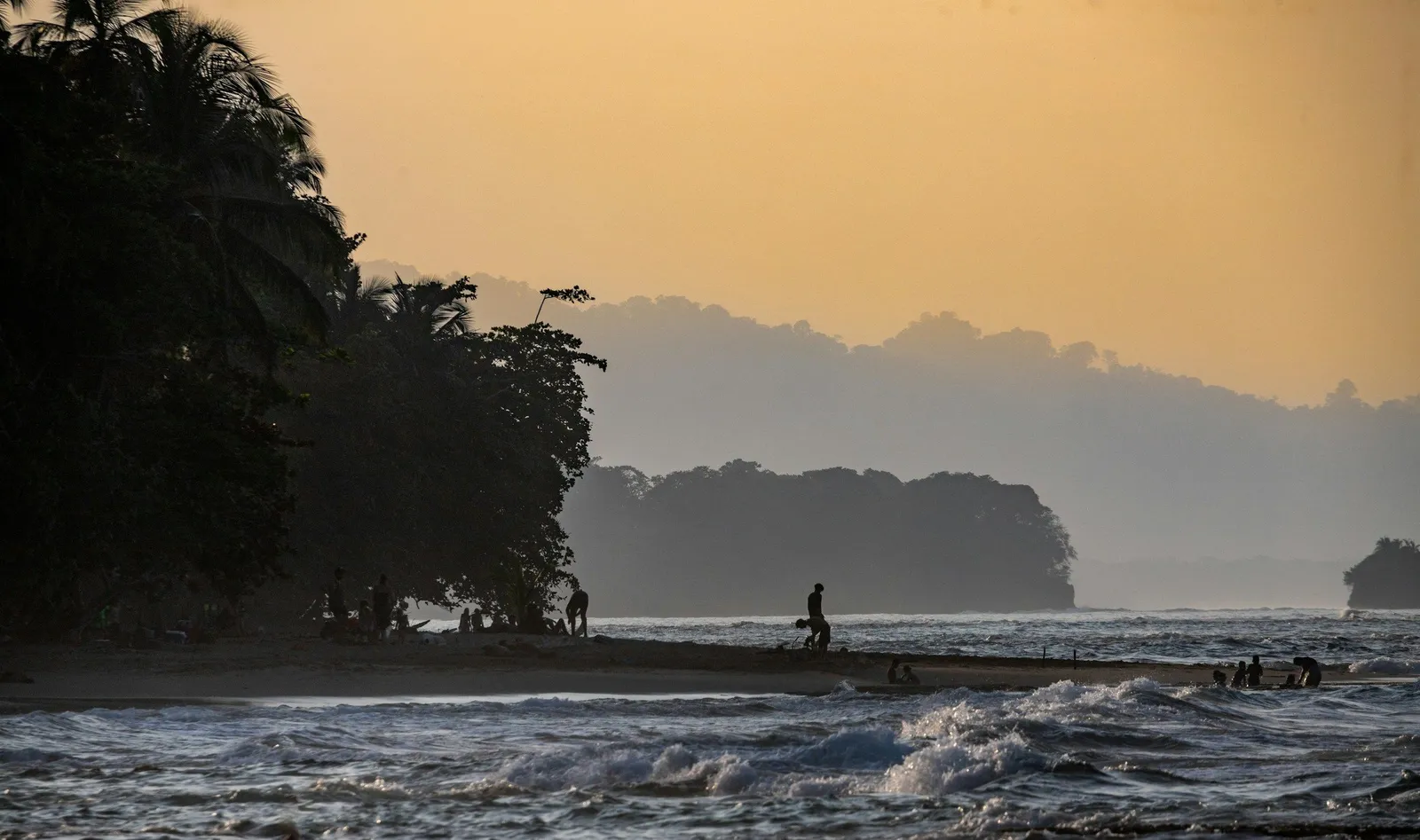 Tropical beach at golden hour with silhouetted palm trees and people swimming, gentle waves and misty jungle headlands in Costa Rica.