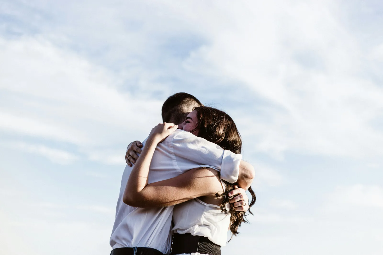 A couple shares a close embrace under a soft blue sky, both wearing white shirts.