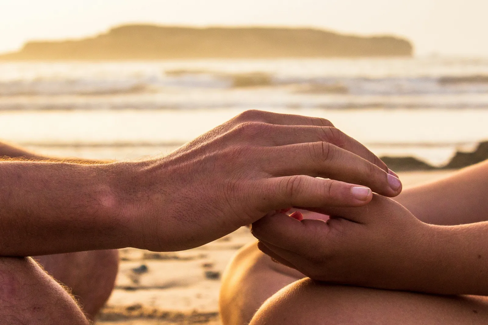 Close-up of two hands holding each other at the wrist against an ocean horizon at sunset.