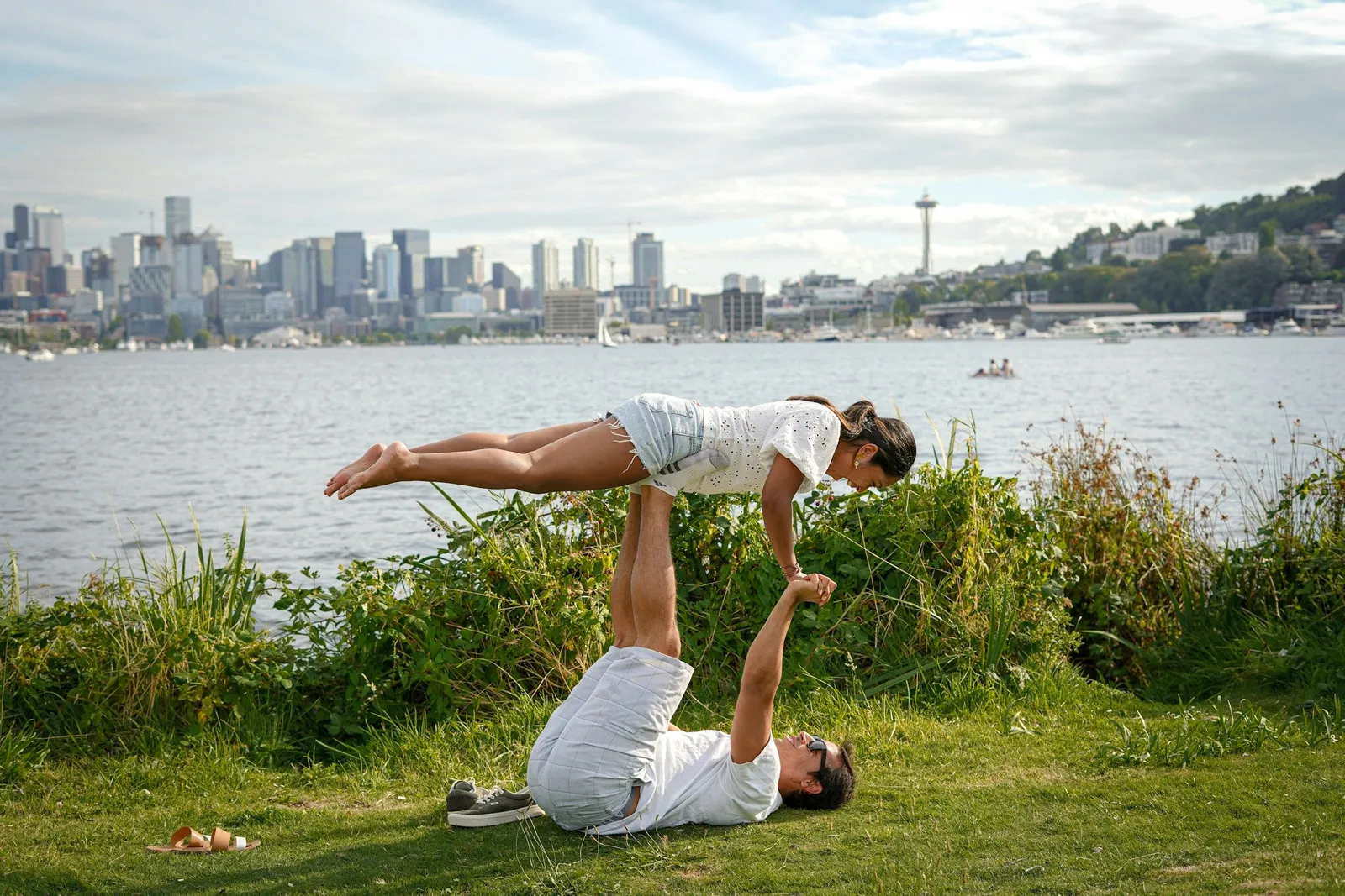 Couple practicing acroyoga on grass with a city skyline and lake in the background, man lying on his back balancing his partner above him in plank pose.