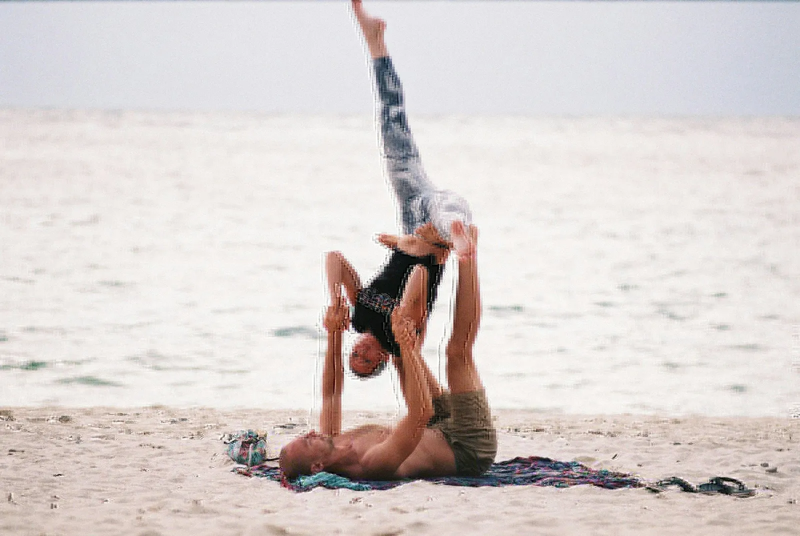 Couple doing acro-yoga on the beach, one person balanced inverted above the other who lies on a towel at the water's edge.