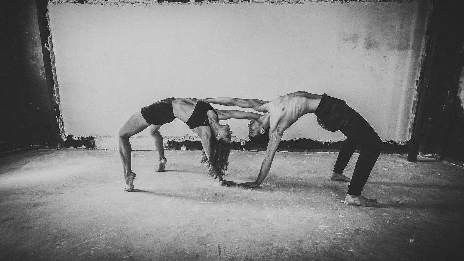 Black-and-white photo of a couple in partner acro-yoga, interlocking wheel poses inside a bare concrete room.