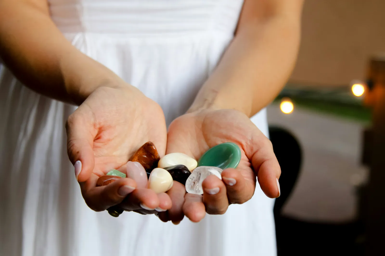 Cupped hands holding polished healing crystals in greens, whites, and clear quartz against a soft warm background.