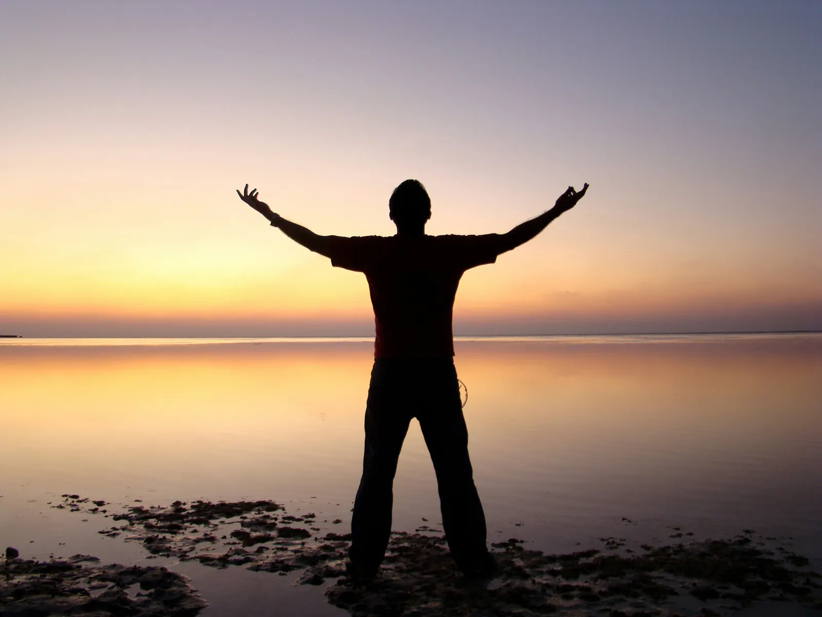 Silhouette of a man standing on a beach at sunset, arms raised wide, ocean behind him, pebbles at his feet.