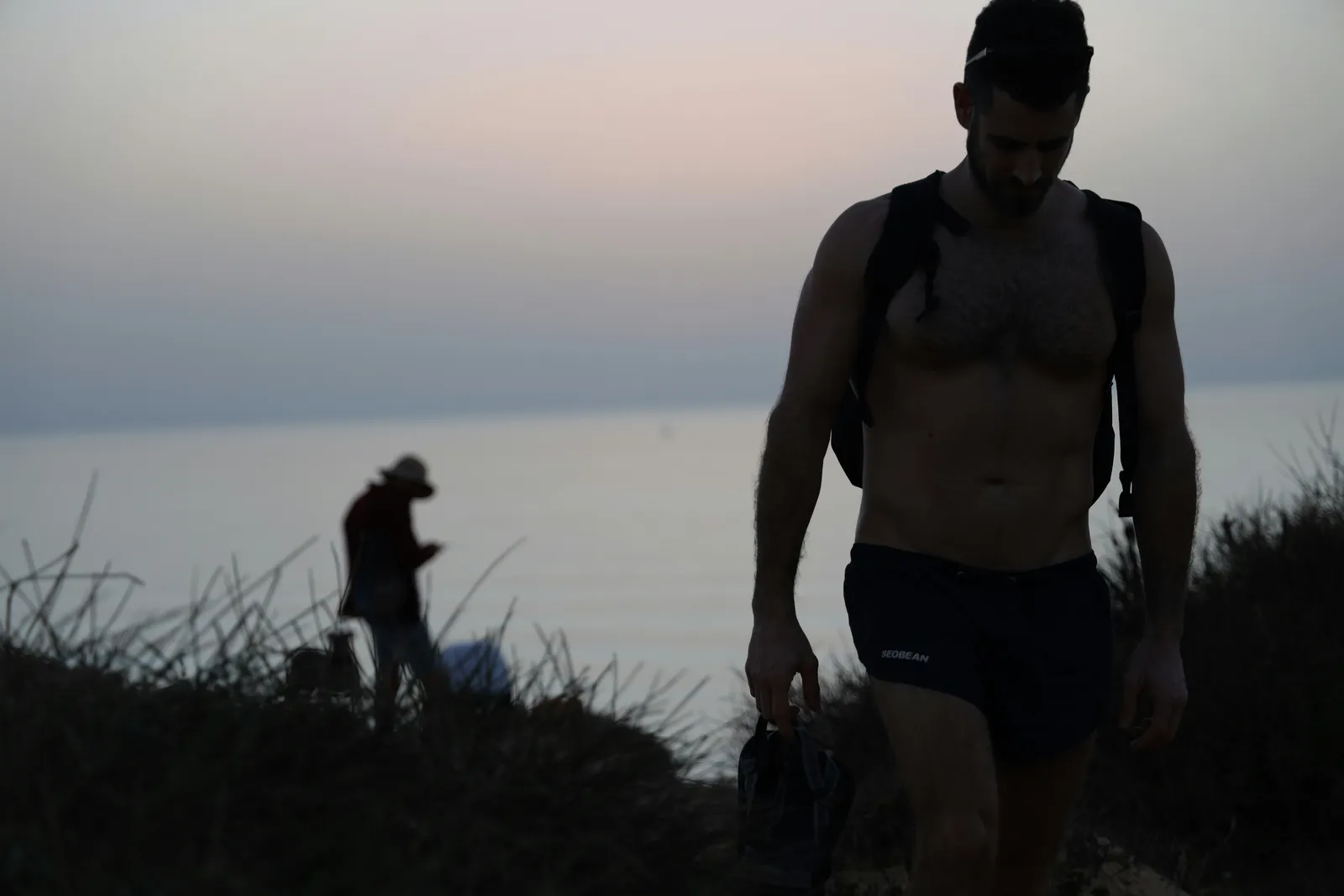 A shirtless man with a backpack walking up a coastal hillside path at dusk, moody sky, distant calm sea behind him.