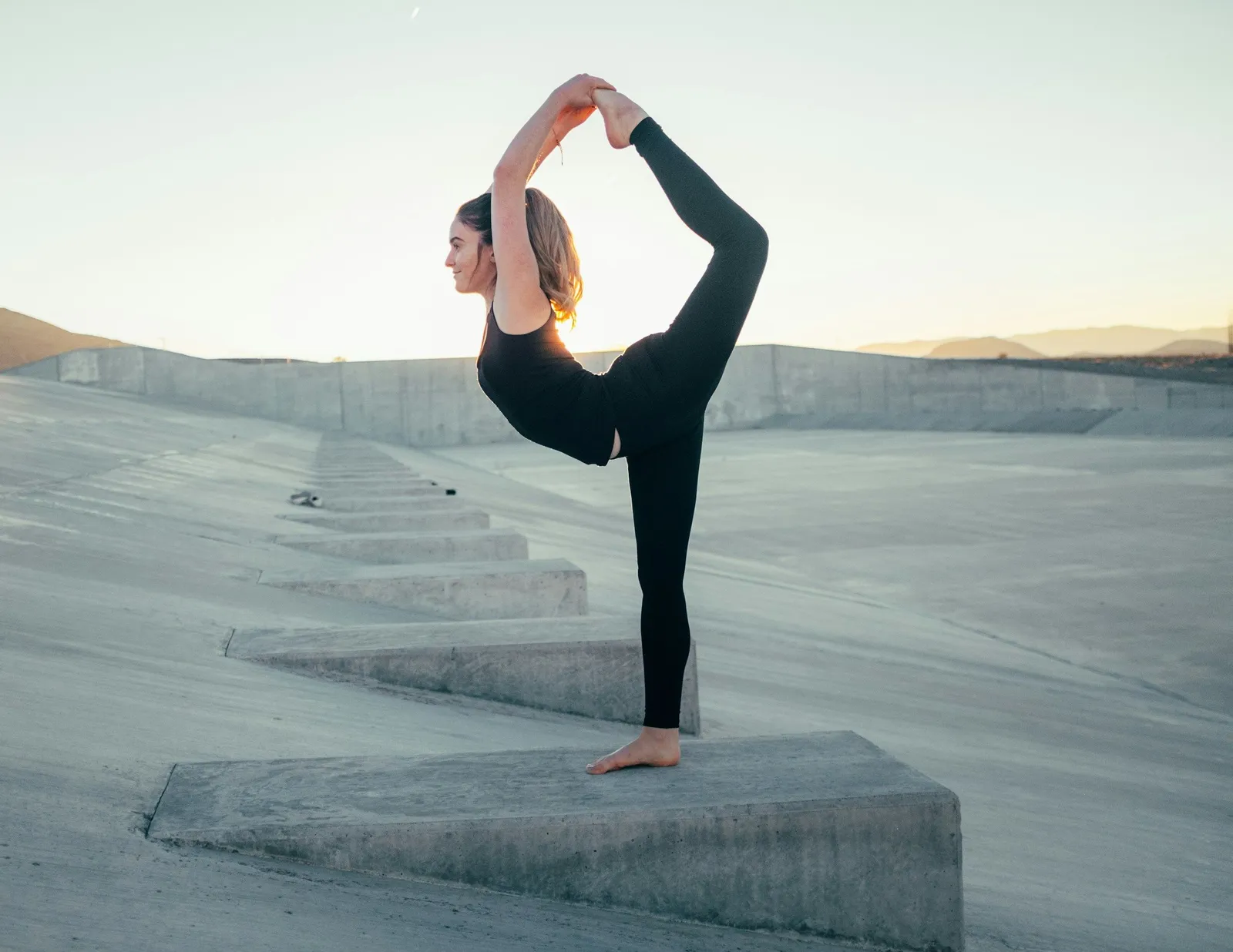 Woman in standing bow yoga pose balanced on one leg on a concrete step at dusk, city background out of focus