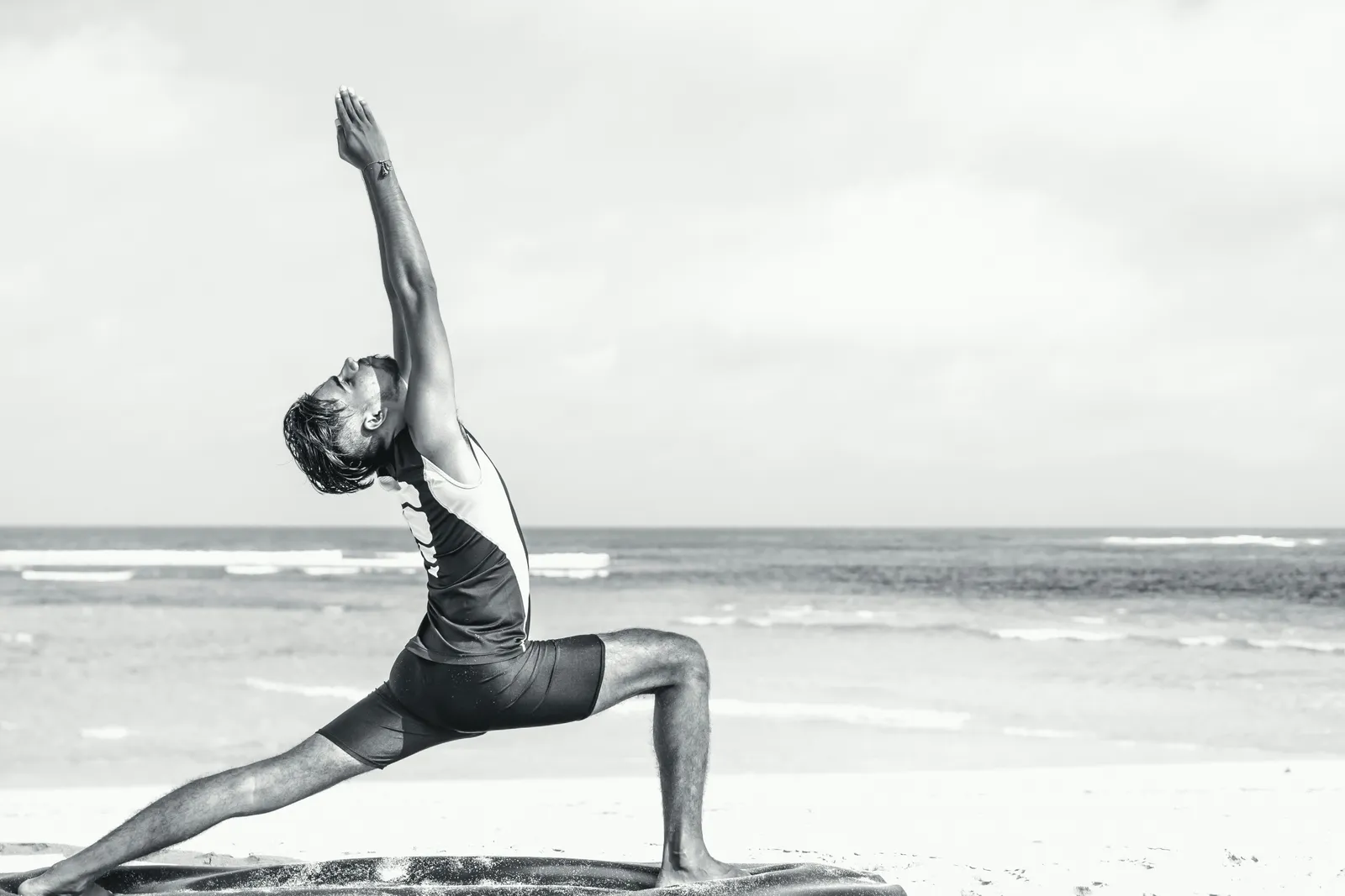 Man in warrior pose with arms raised on a sandy beach, black-and-white photograph, ocean in background