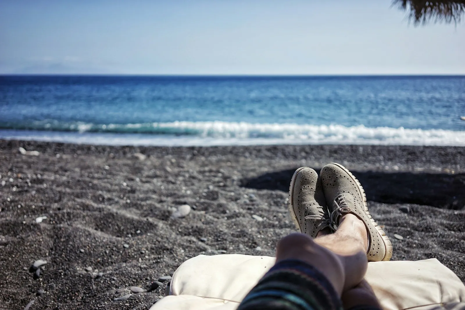 First-person view of crossed feet in suede shoes resting on a cushion on a black pebble beach with turquoise sea ahead.