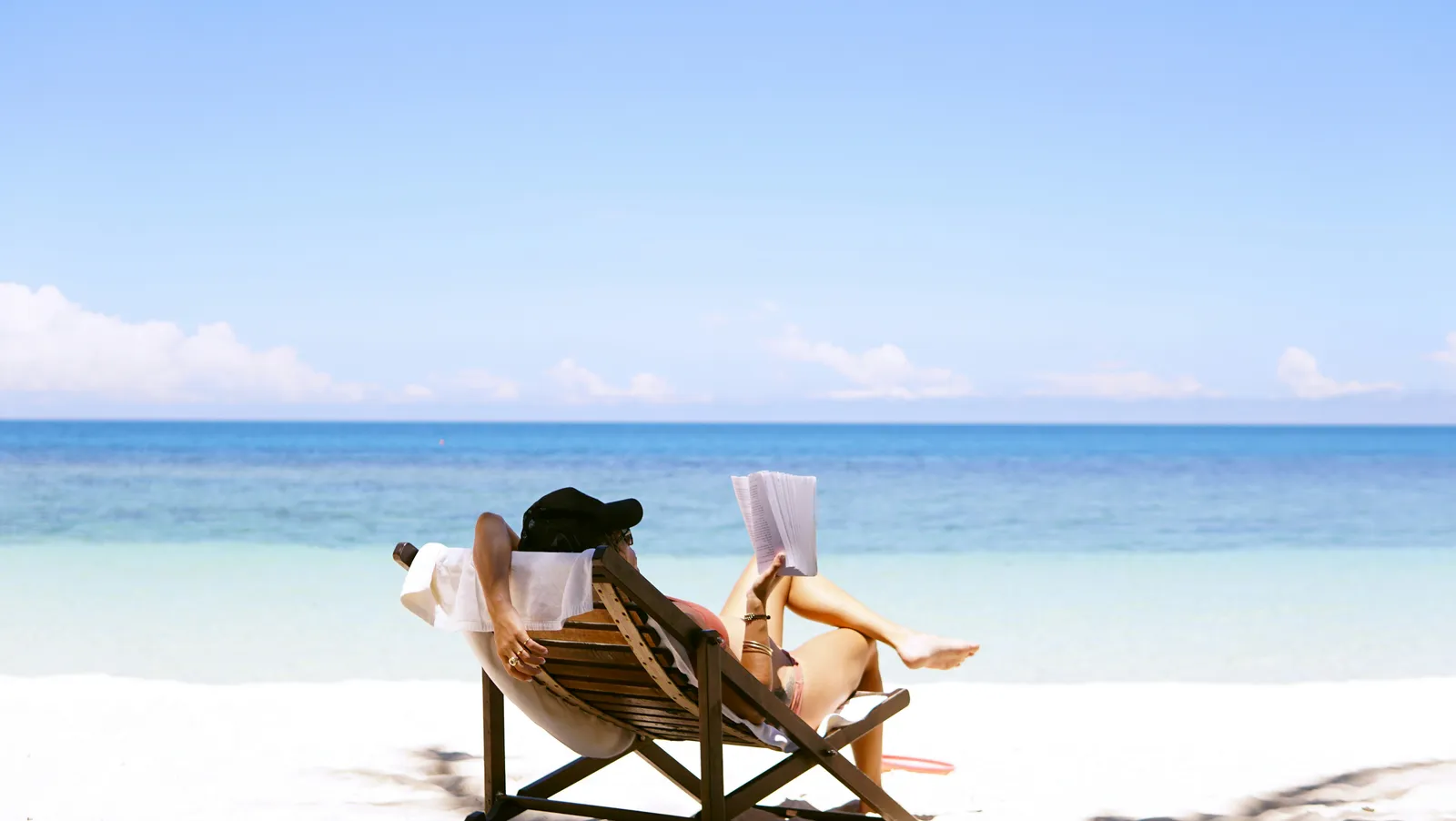 Woman in a sunhat reclining on a wooden beach chair reading a book on white sand near the water.