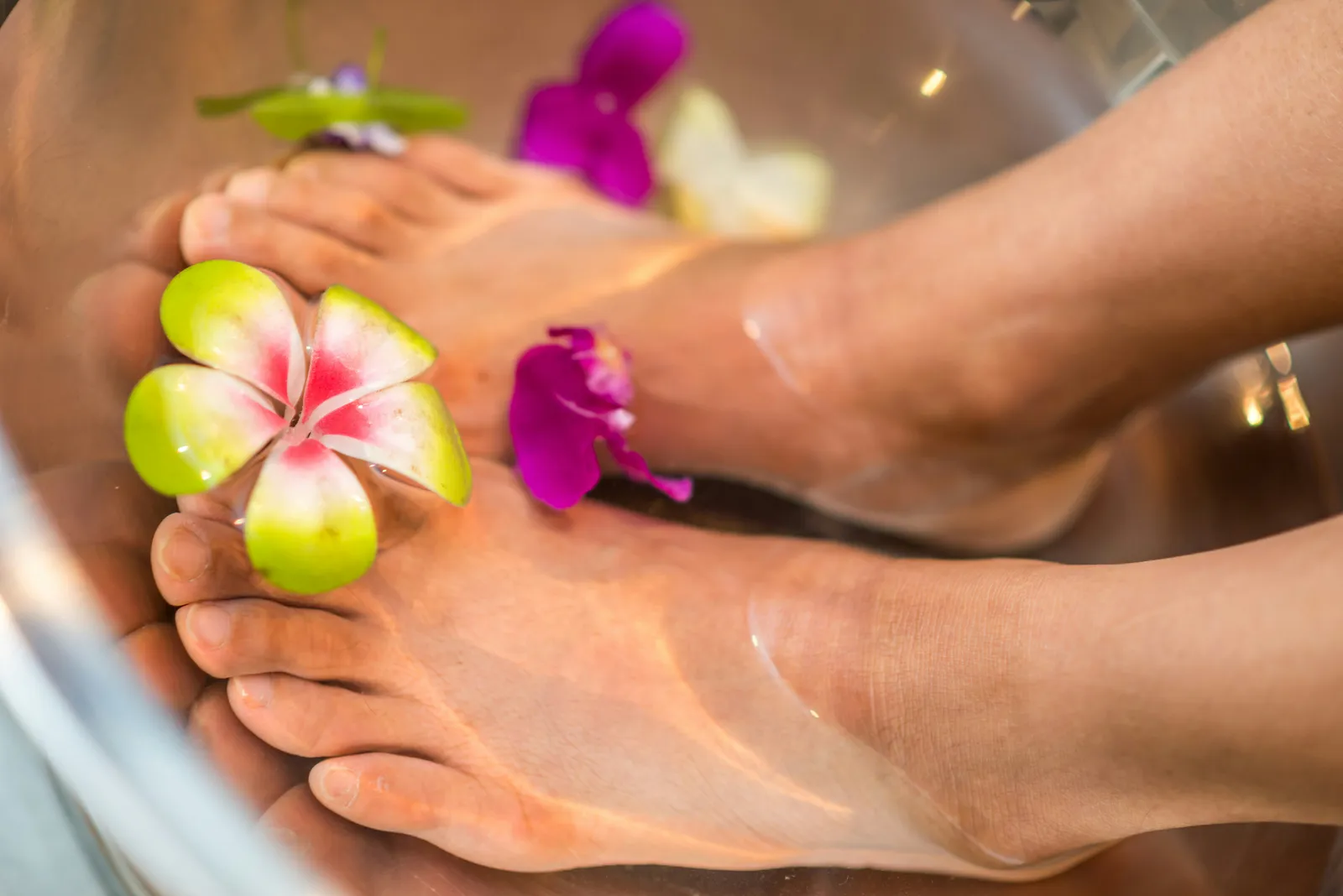 Two pairs of feet rest in a metallic bowl of water with floating frangipani and orchid flowers during a spa foot soak. Close-up, warm tropical spa mood.