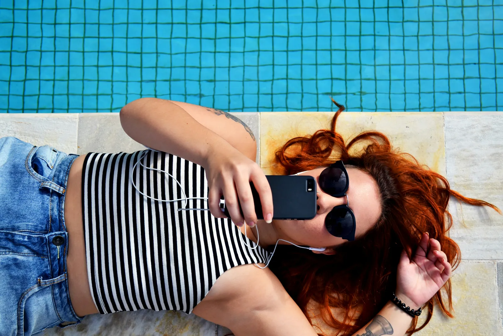 Overhead view of a red-haired woman lying poolside on a striped towel taking a selfie with her phone while wearing sunglasses.