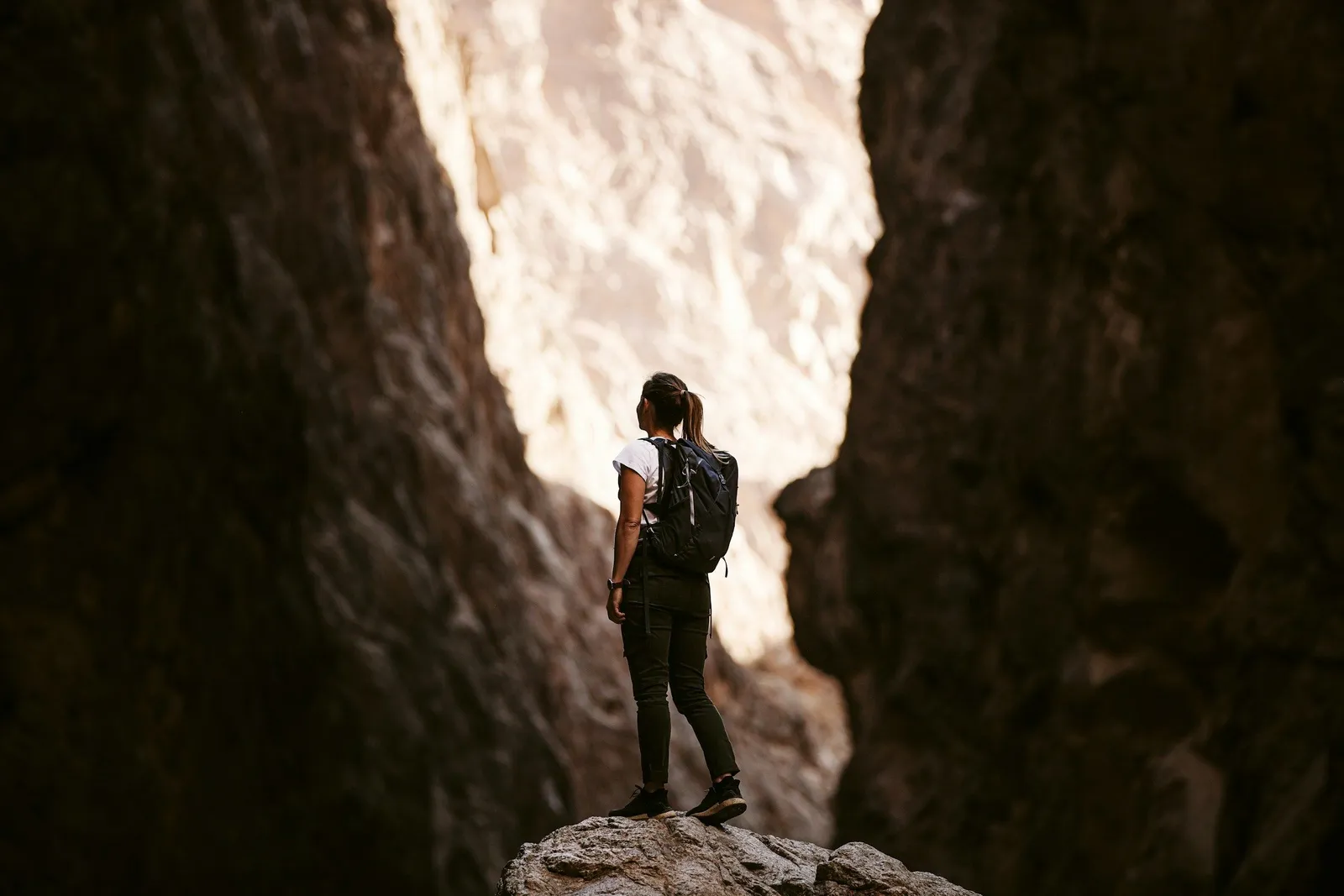 Woman with backpack standing on a rock inside a narrow rocky gorge looking up toward sunlit canyon walls.