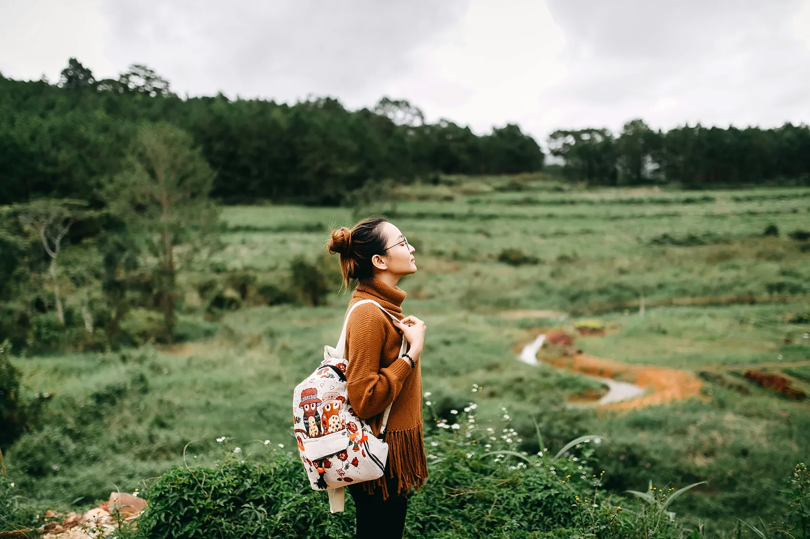 A young woman with a backpack stands on a hillside path looking out over rolling green countryside under a cloudy sky. Reflective traveler mood.