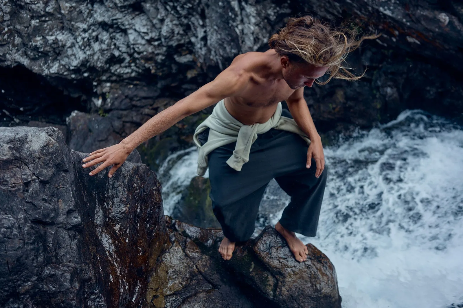 Shirtless long-haired man in loose pants balancing barefoot on wet rocks beside a rushing waterfall.