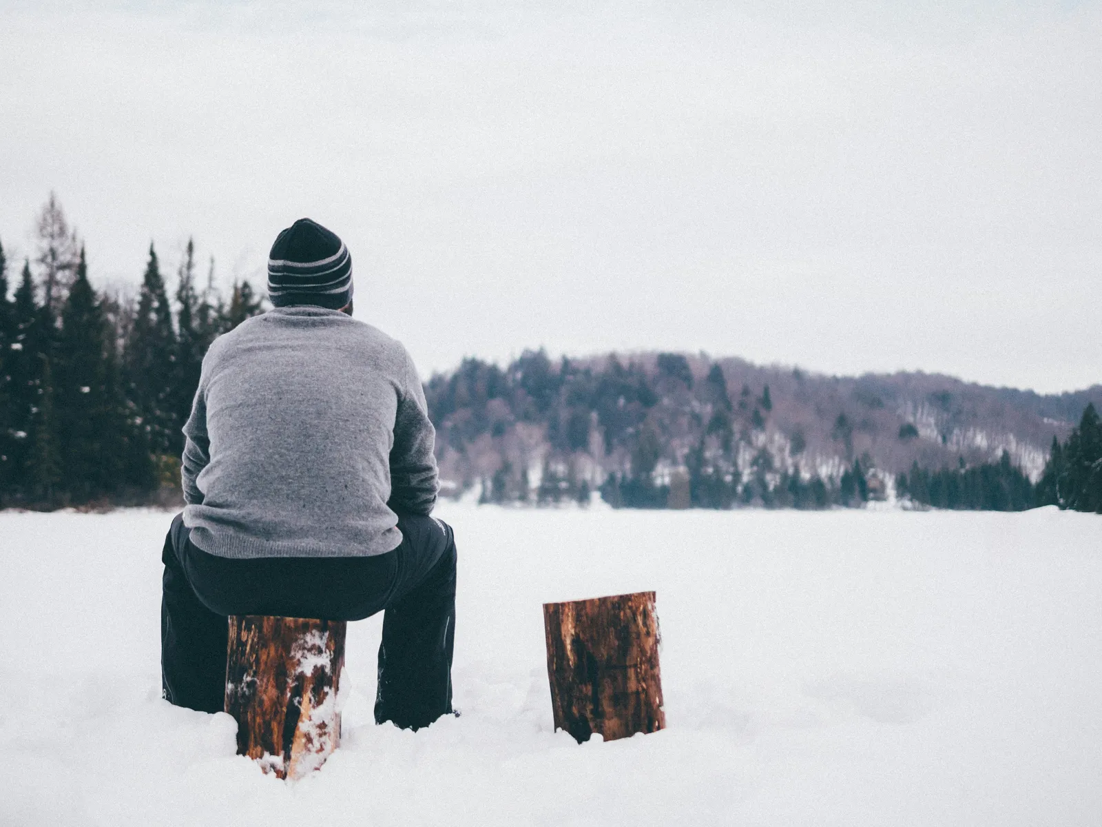 Person in a beanie and gray sweater sitting alone on a snow-covered frozen lake, facing a distant winter forest.