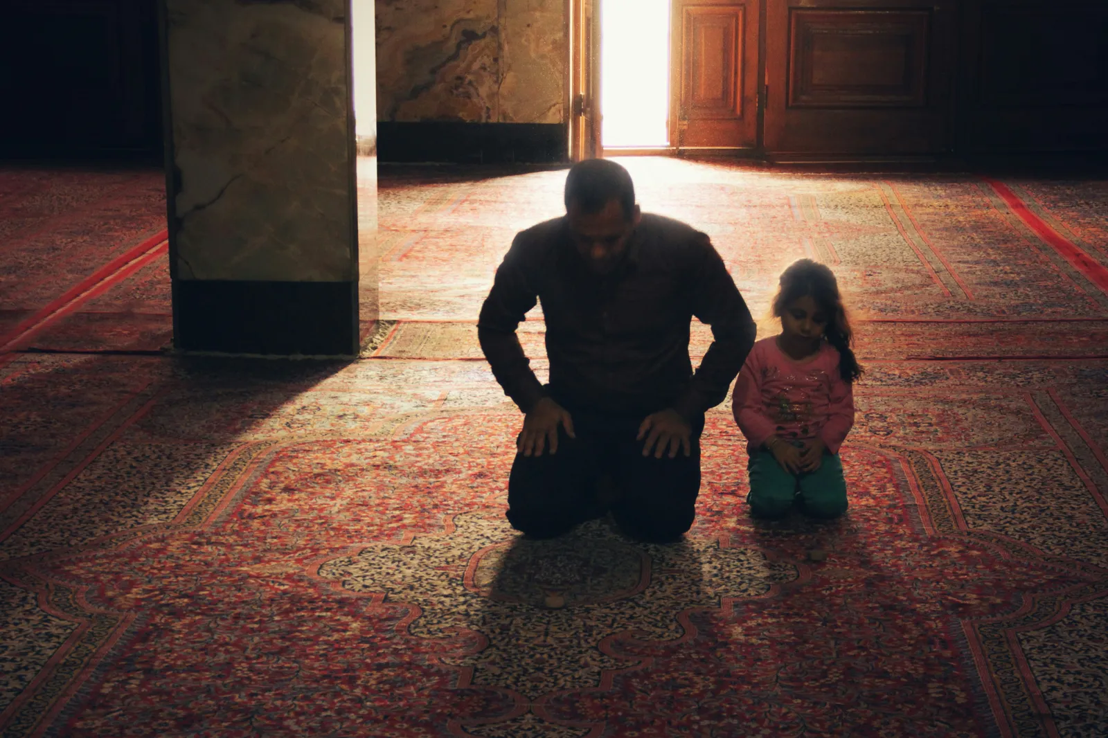 A man and a young girl kneel in prayer on an ornate red Persian carpet inside a dimly lit room with sunlight streaming through a doorway. Sacred, intimate mood.