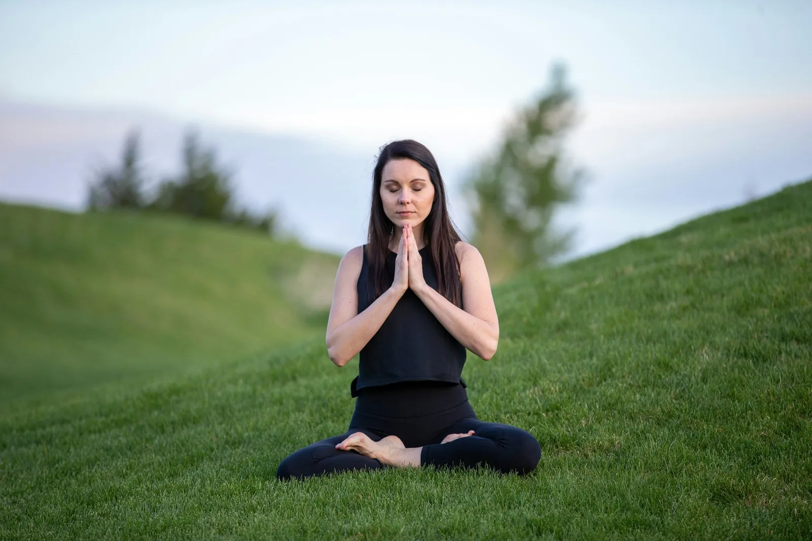 A dark-haired woman meditates in lotus pose on bright green grass on a hillside, eyes closed, hands in prayer. Fresh outdoor meditation scene.