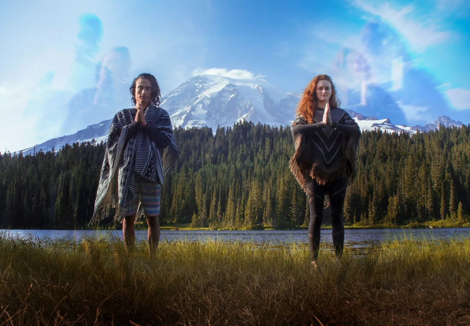 Two long-haired people in shawls standing in prayer pose by a mountain lake with Mount Rainier and forest behind, soft light surrounding them.