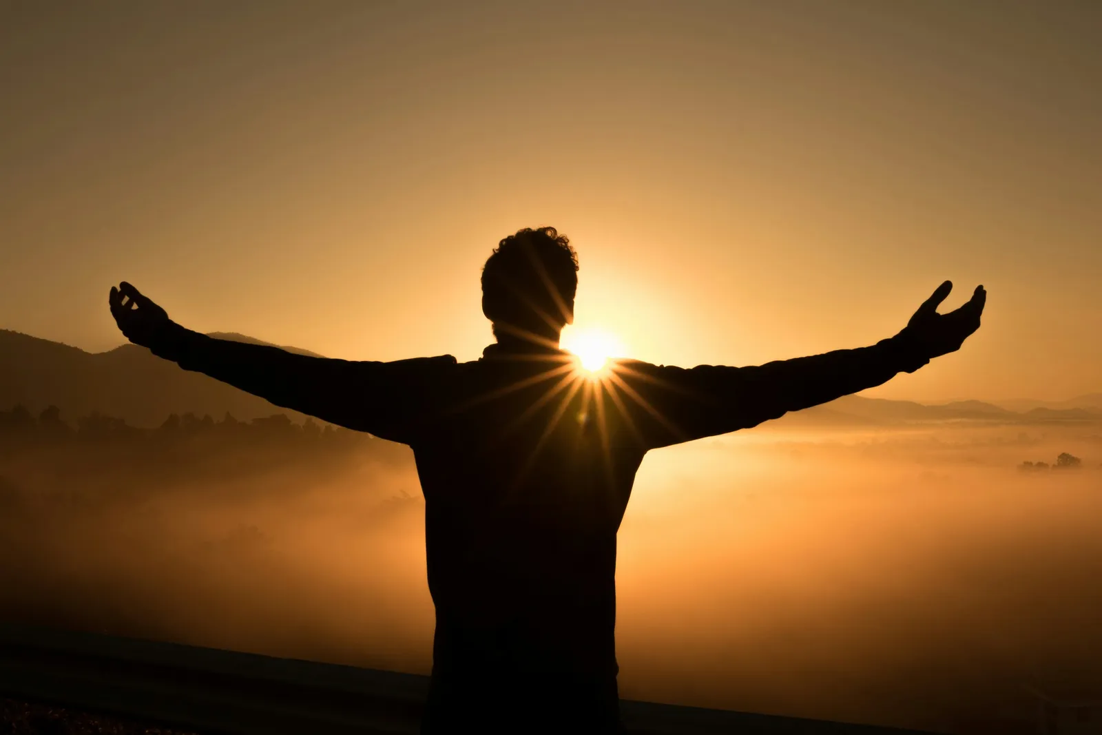 Silhouette of a man with arms outstretched in a victory pose facing a glowing sunrise over misty mountains, an image of the open and uplifted feeling that often follows inner retreat work.