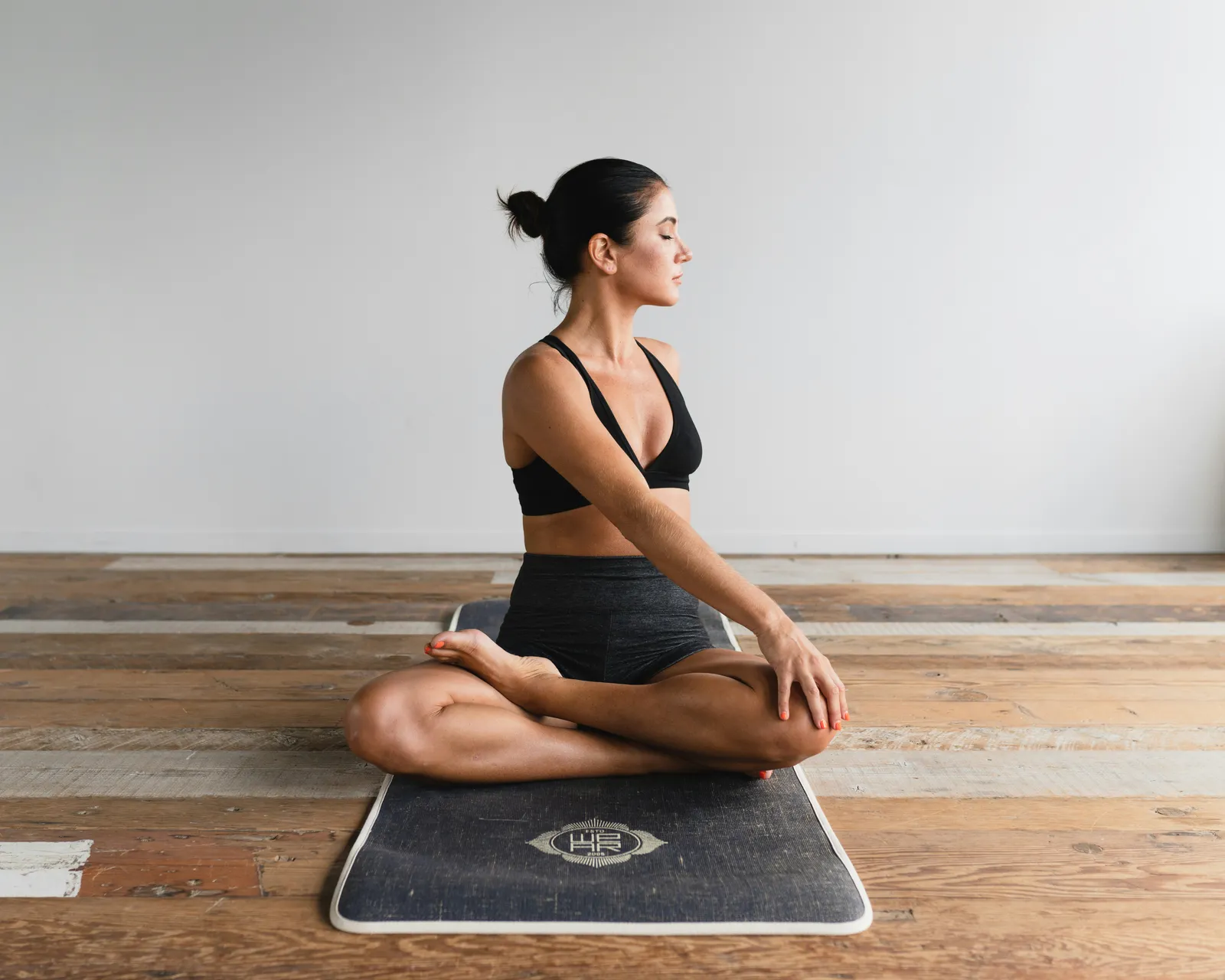 Woman in black sports bra seated in lotus pose with eyes closed on a yoga mat over a wooden floor. Quiet indoor meditation.