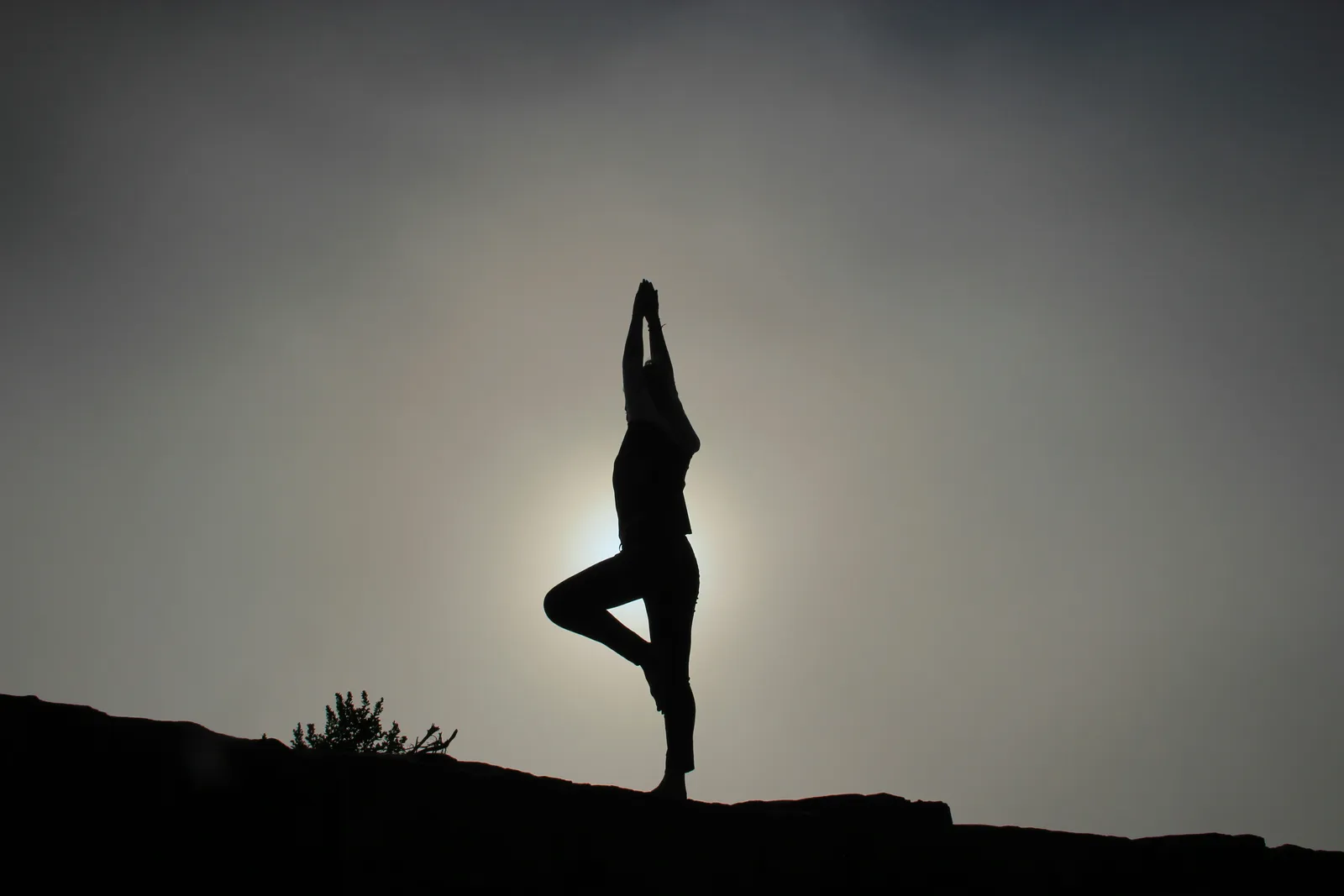 Silhouette of a person in tree pose on a rocky ridge backlit by a hazy sun in a gray sky. Balance and stillness in an outdoor setting.