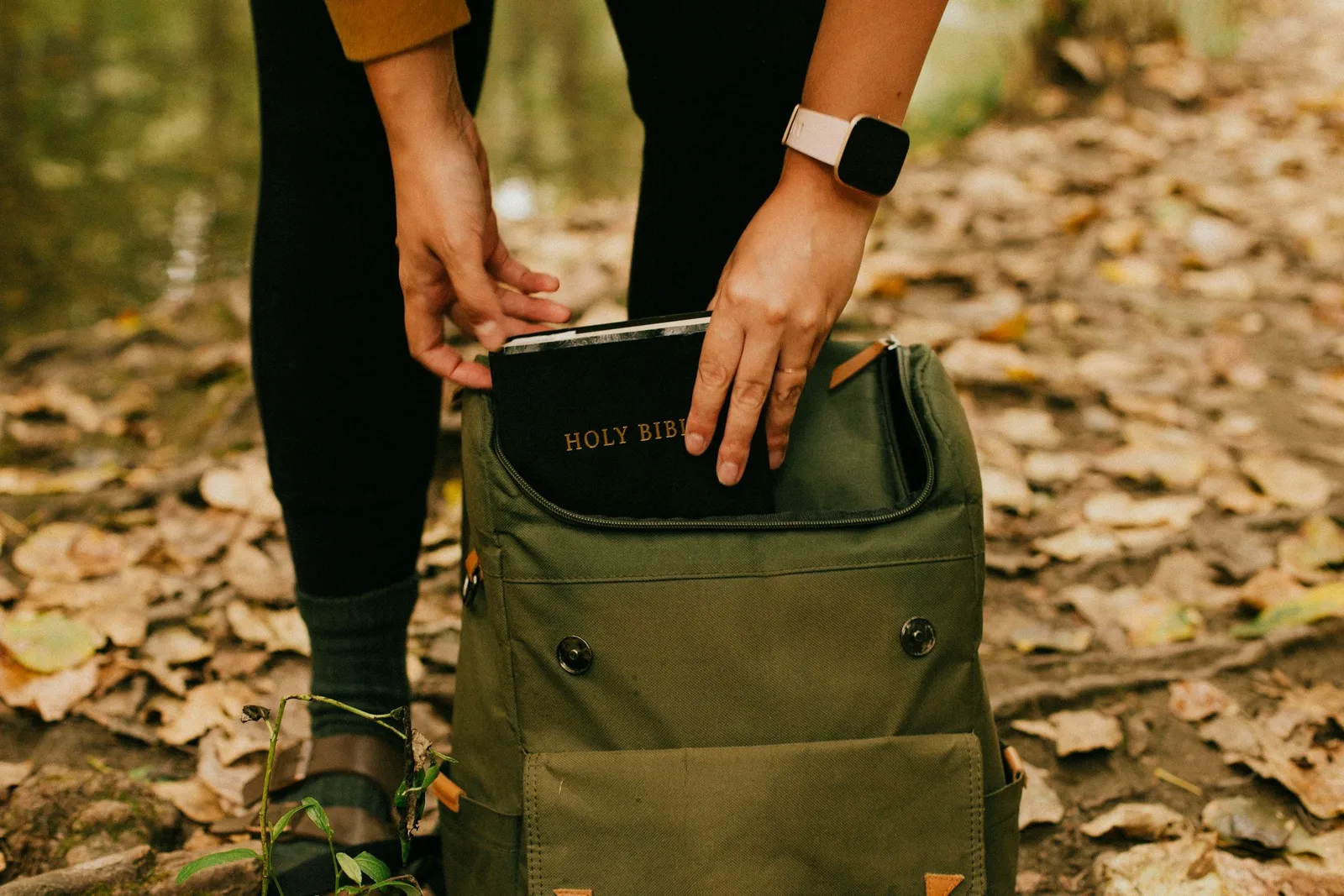 Person placing a book into a backpack among autumn leaves, preparing for a spiritual retreat.