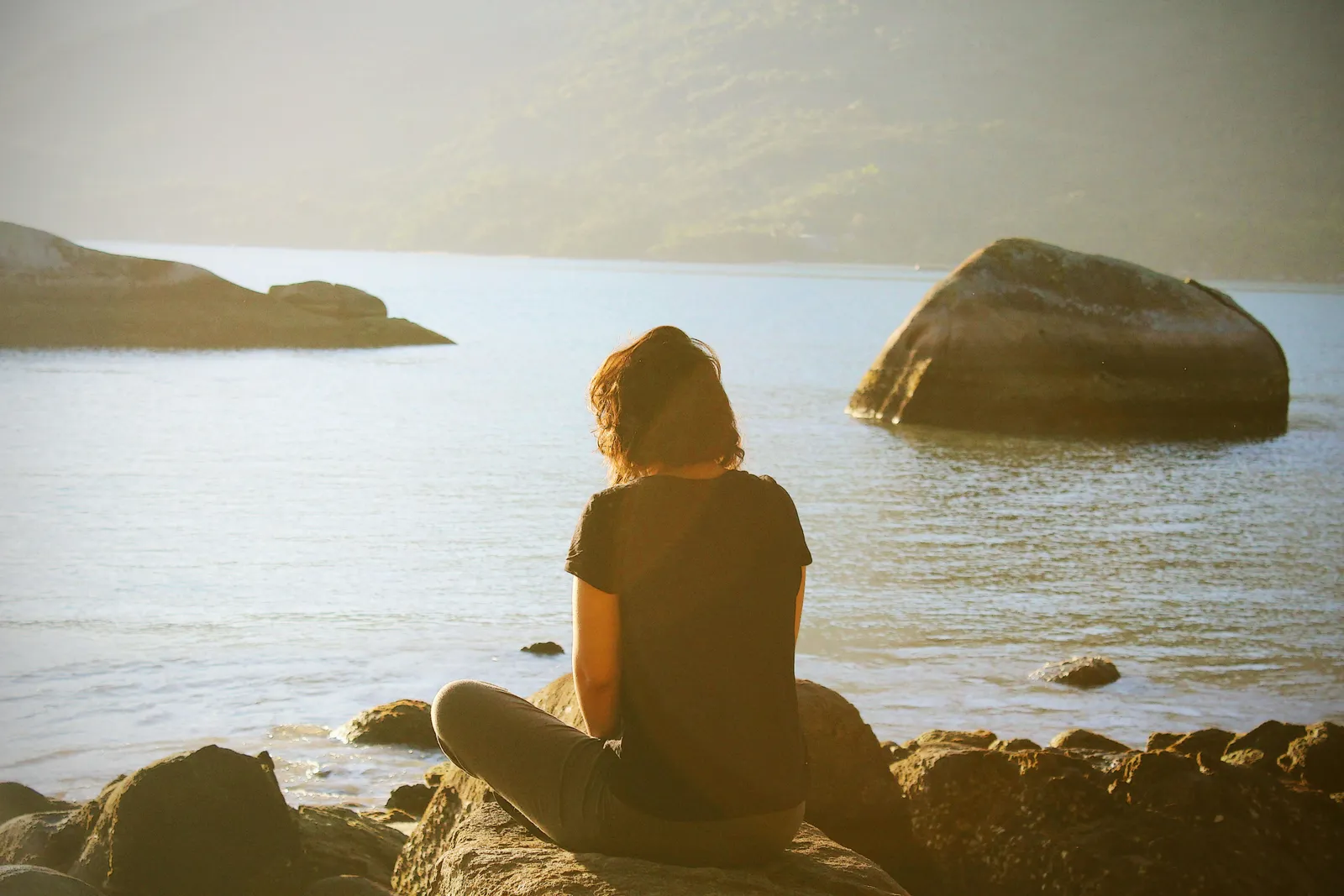 A woman in a dark t-shirt sits on rocks at the edge of a misty lake gazing toward distant green mountains in warm golden light.
