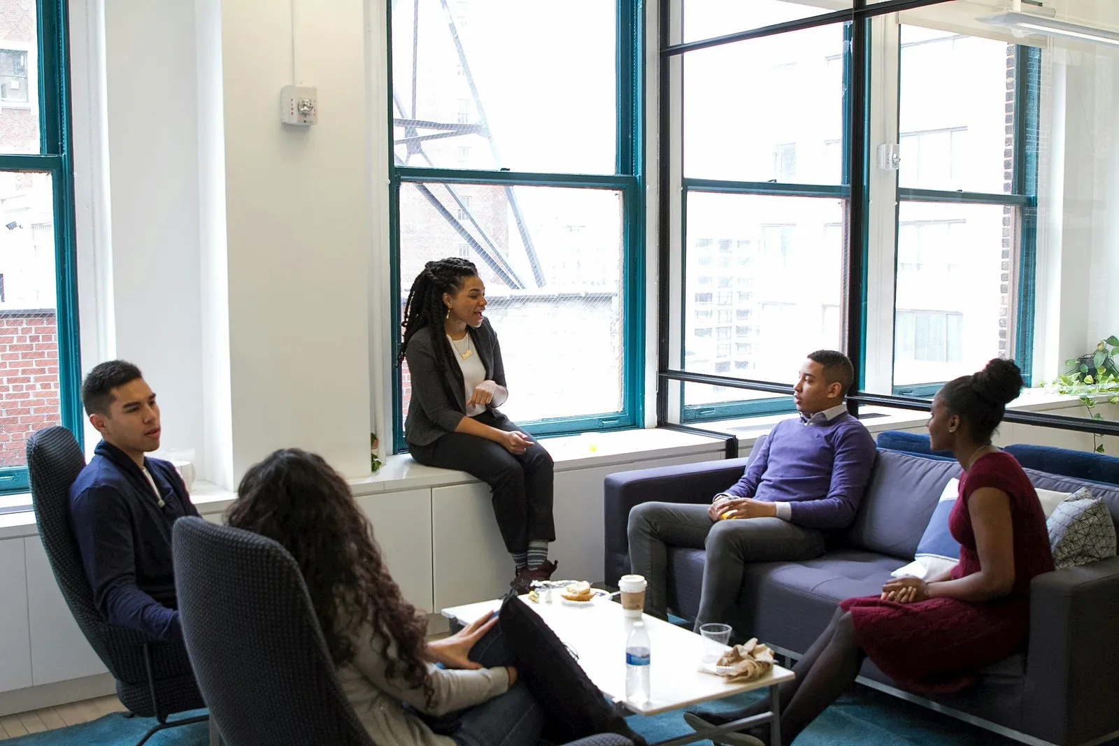 Diverse group of professionals having a casual meeting in a bright office lounge with one person seated on a window ledge.