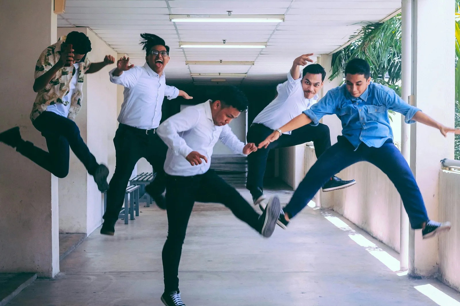 Five young men in white shirts and dark trousers leaping into the air in a bright hallway, energetic and playful.