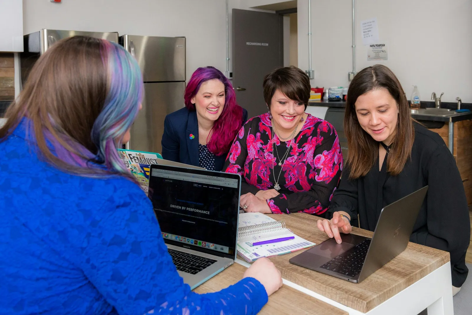 Four women around a wooden table with two laptops collaborate happily in a bright office kitchen. Casual, energetic team workshop vibe.
