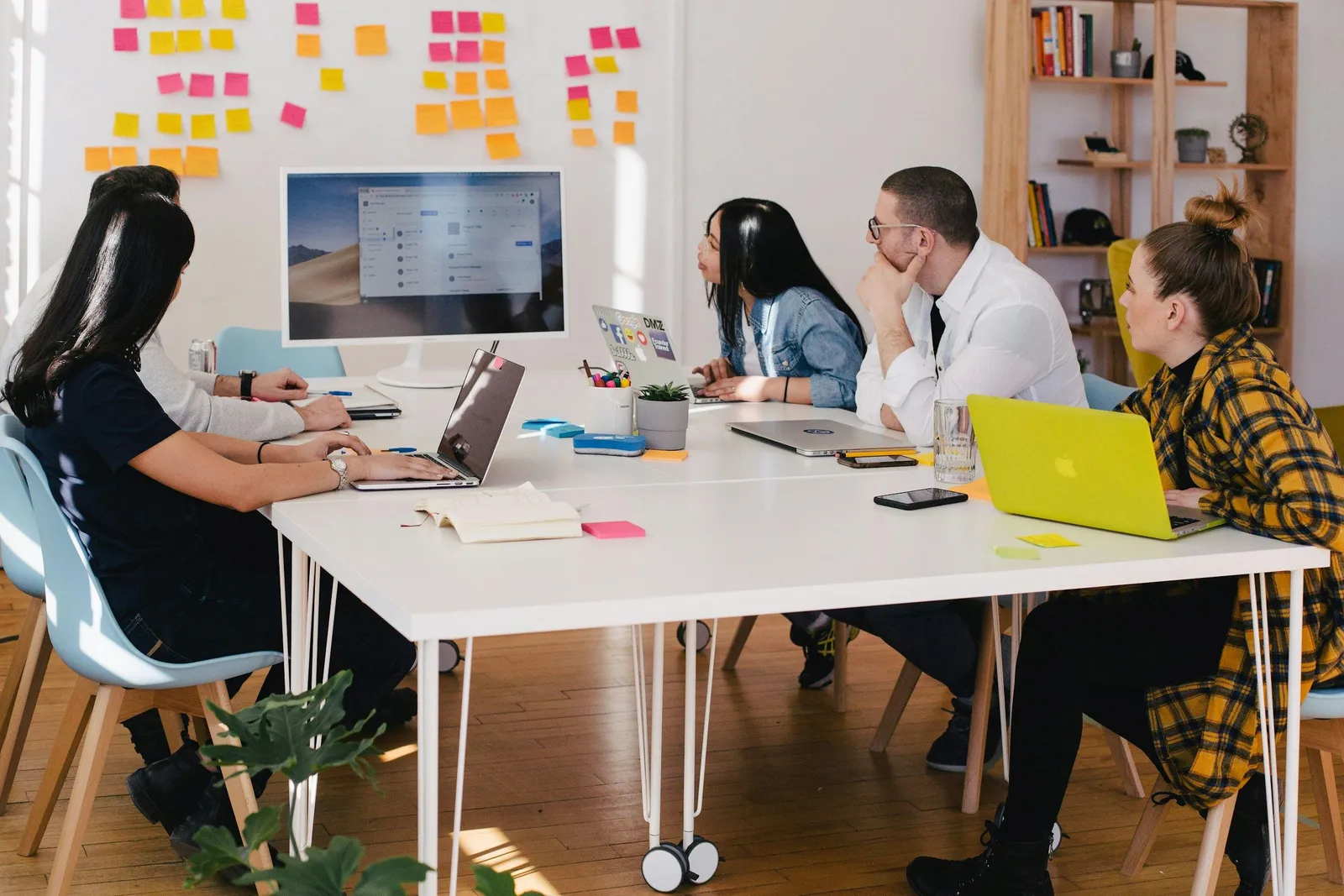Five people work around a white table with laptops and notebooks in a bright office covered in colorful sticky notes on the wall. Team brainstorming meeting.