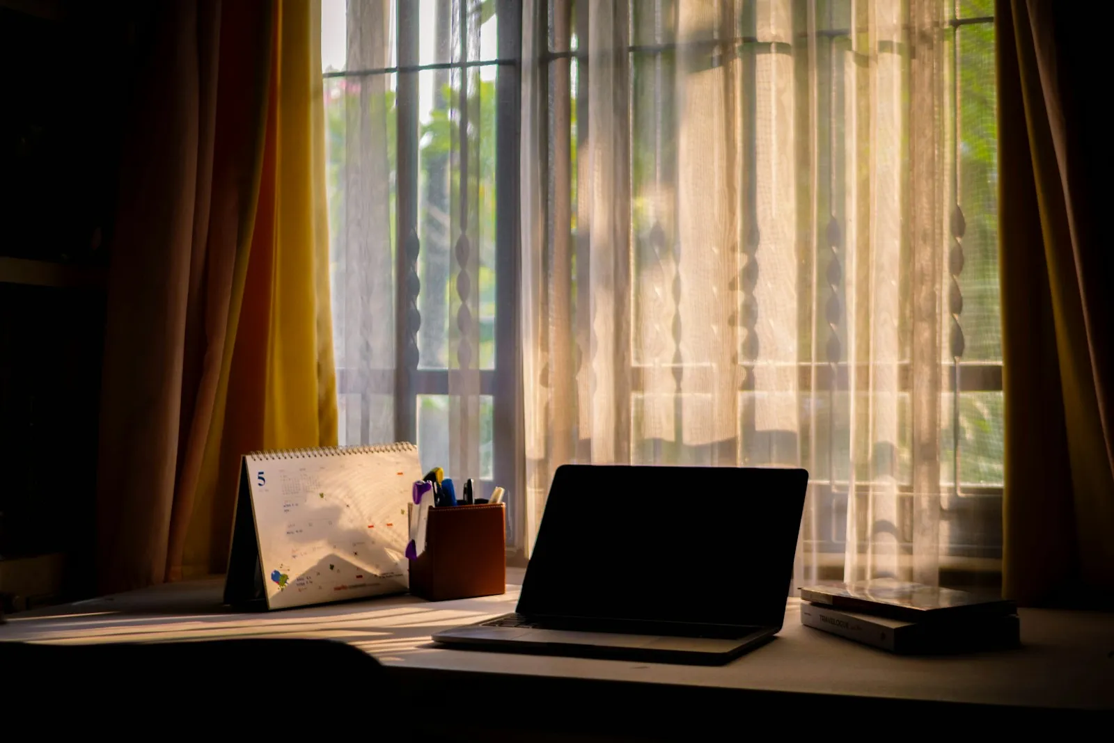 Open laptop next to a desk calendar and a pen holder on a desk in front of a sunny window with sheer curtains.