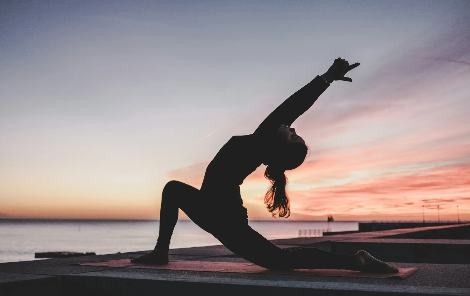 A woman in a low crescent lunge on a seaside terrace at sunset, the kind of practice that's easy on retreat and harder to keep up at home.