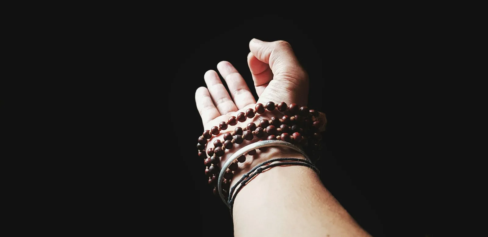 Single open hand emerging from black background wearing wooden mala prayer beads. High-contrast, minimalist.