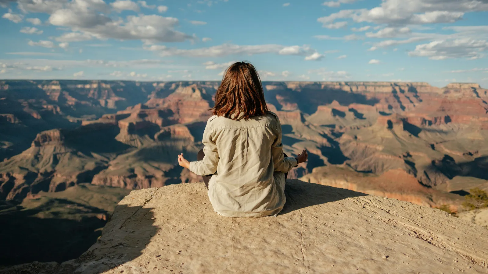 Woman seated cross-legged on a rocky ledge above the Grand Canyon, eyes closed, hands resting on knees, expansive sky and canyon behind her