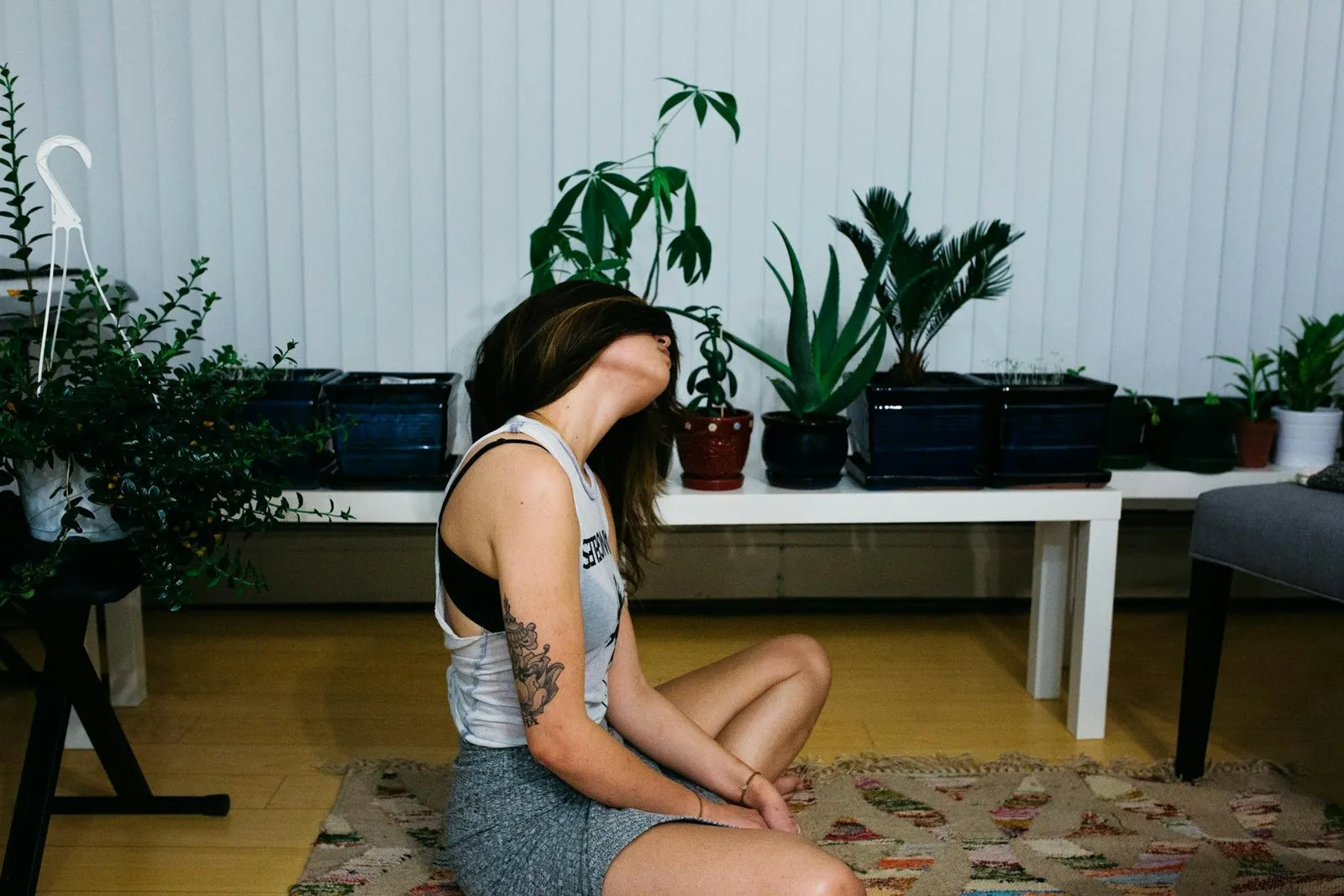 Tattooed woman sitting cross-legged on a rug at home in front of shelves of houseplants, eyes closed in a resting pose.