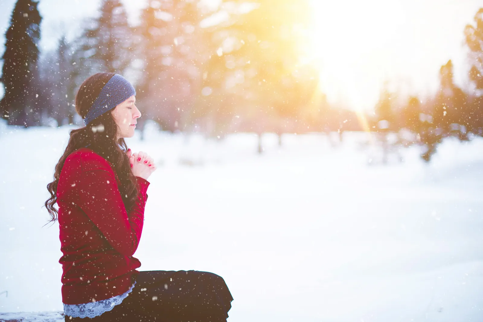 Woman in a winter prayer pose in a snow-covered clearing, warm sunlight breaking through the trees.