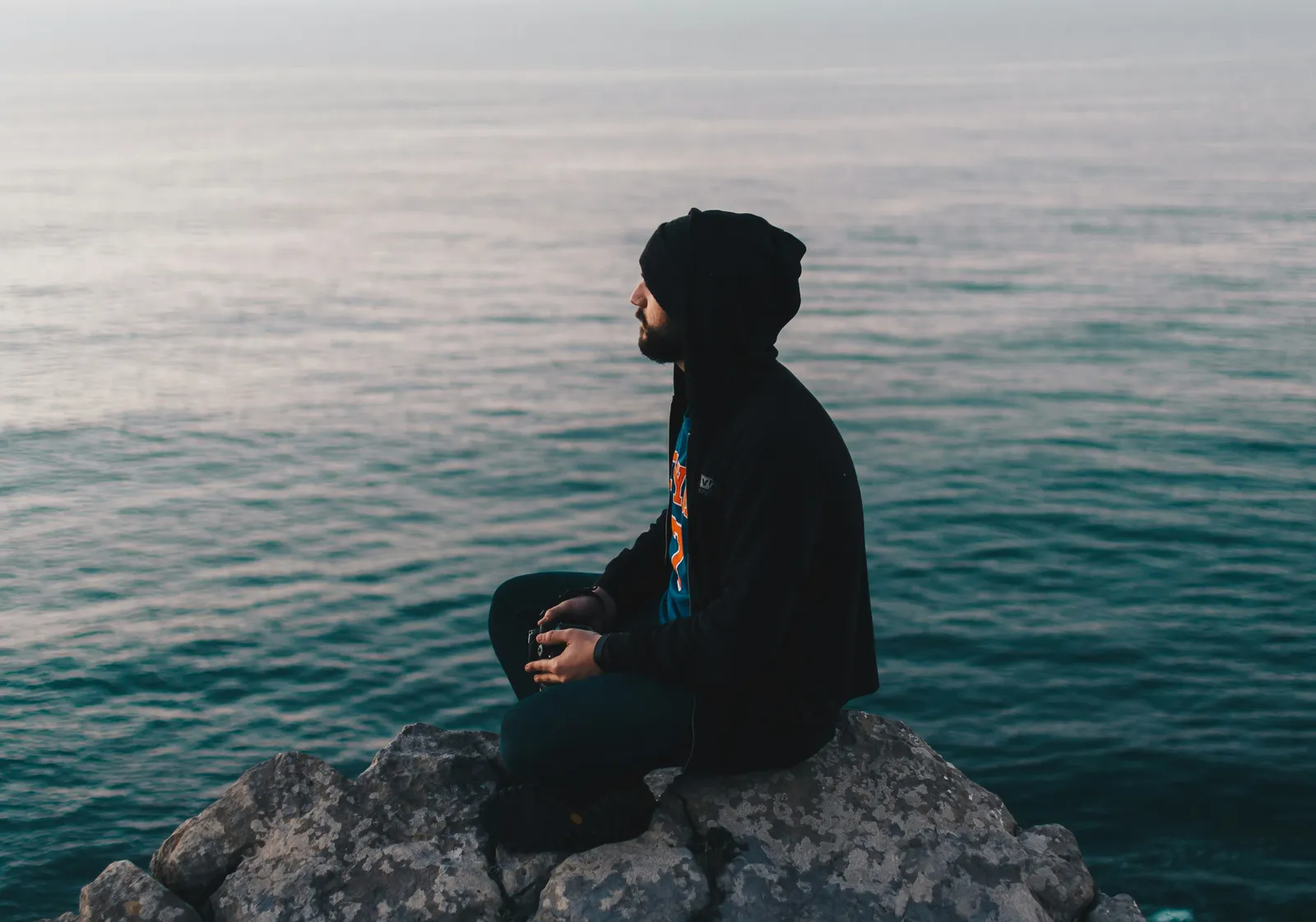 Man sitting cross-legged in meditation on coastal rocks, looking out to sea.