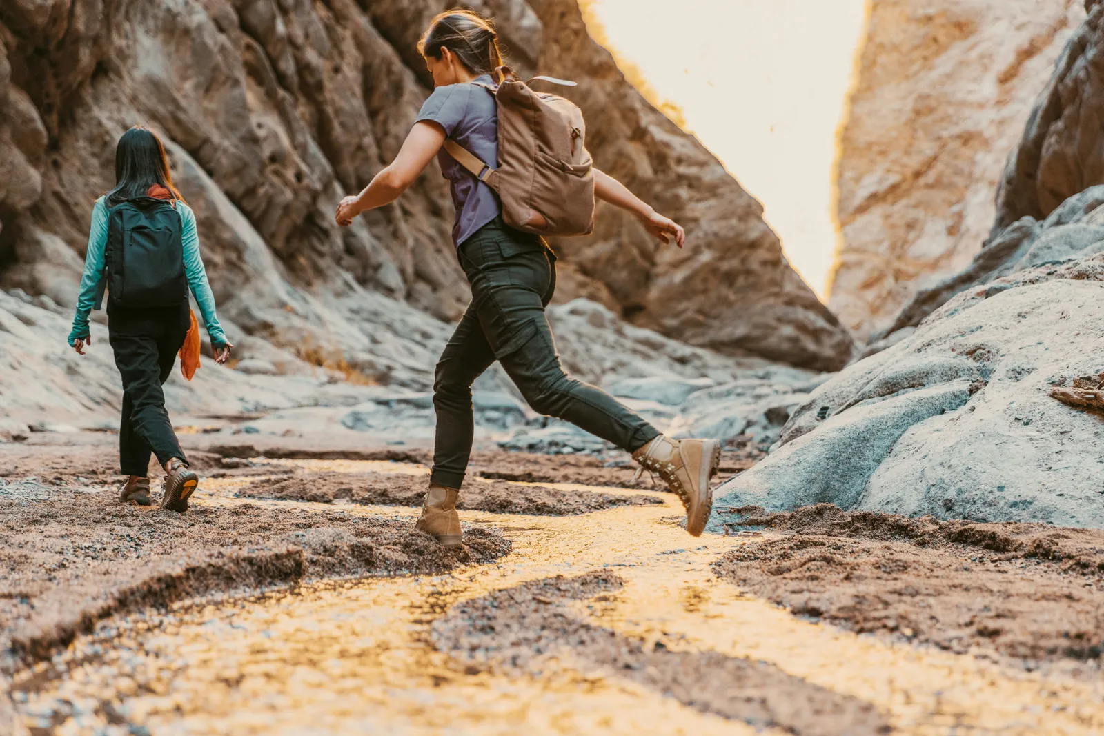Two women with backpacks hiking and jumping across rocks in a sunlit canyon with rugged stone walls around them.