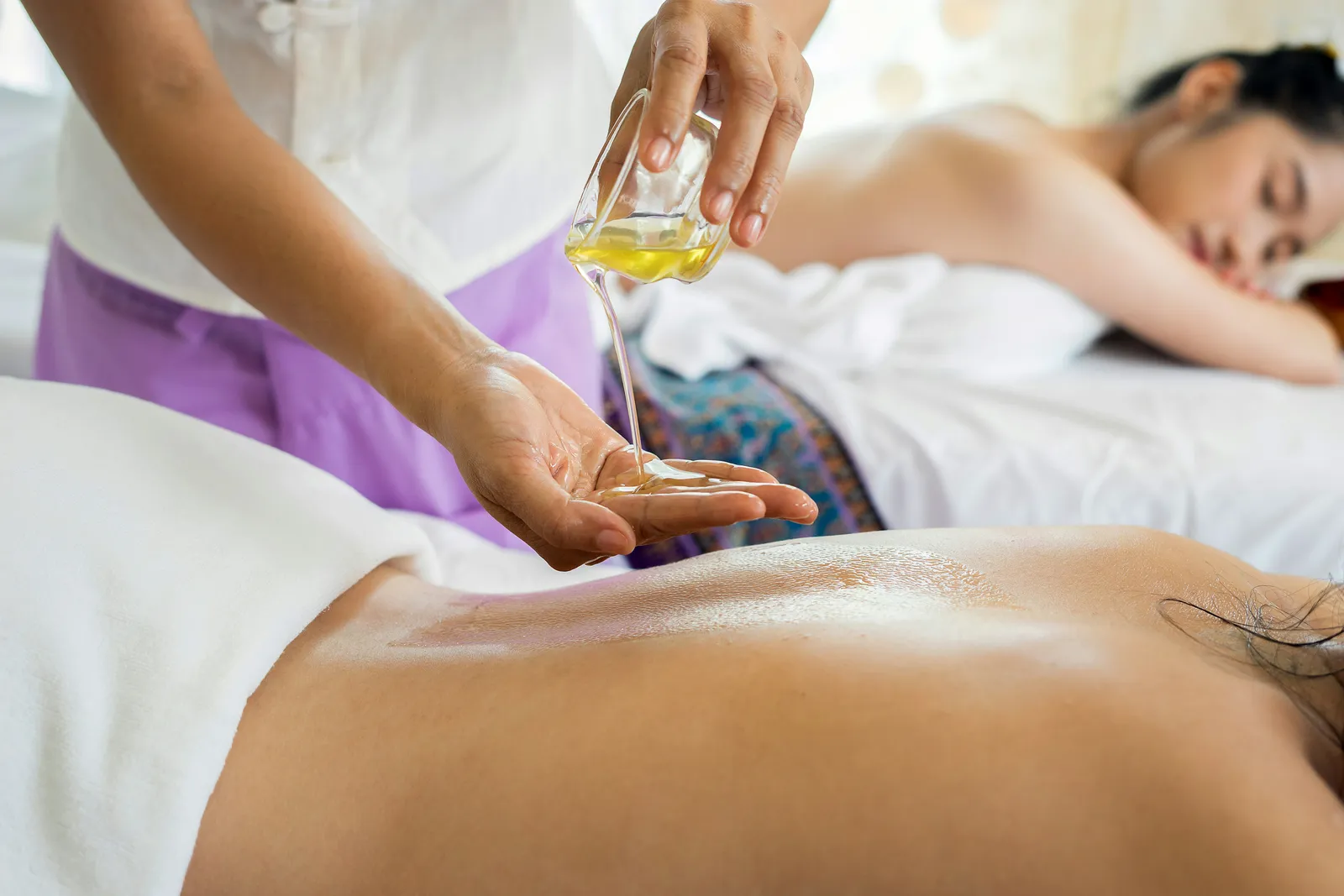 Massage therapist pouring yellow oil into her palm above a client's back during a spa treatment, with another woman resting on a nearby table.