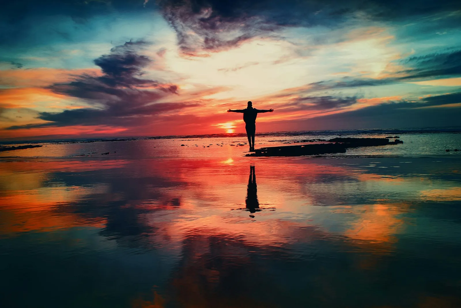 Silhouette of a person standing on wet sand at sunset with arms outstretched, the vivid sky reflected in the shallow water below.