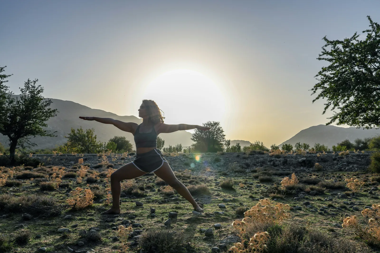 Woman in warrior II pose outdoors at sunset with mountains behind. Mountain and forest settings are one of four main formats for beginner programs.